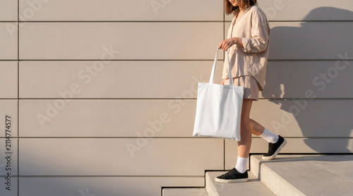 Blank white tote bag mockup held by a woman walking down modern stairs with large copy space