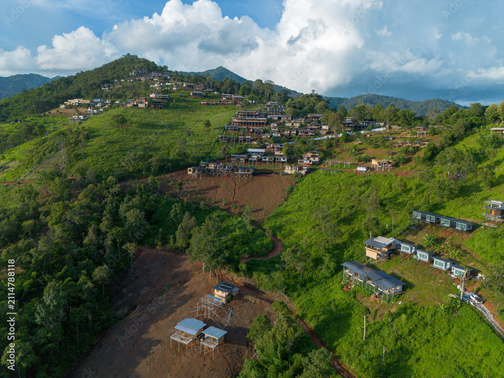 Fototapeta premium Hillside village with terraced houses and green vegetation under dramatic clouds, peaceful rural landscape with winding paths and scattered cabins conveying serenity and openness