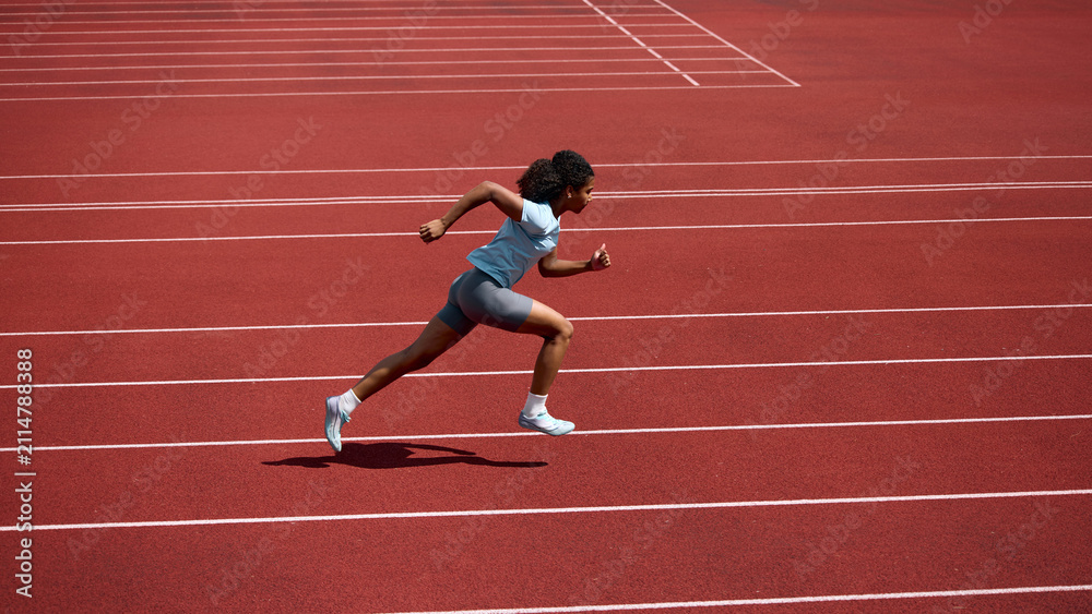 Fototapeta premium Teenage female runner sprinting at full speed across red athletics track. Concept of speed development, track and field sports, summer competition.