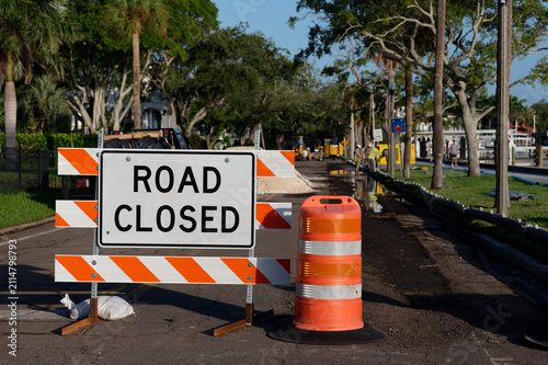 A reflective Orange Road Closed barricade with an orange detour arrow blocks a suburban street, held down by sandbags; sunlight illuminates the warning stripes as traffic is diverted around neighborho