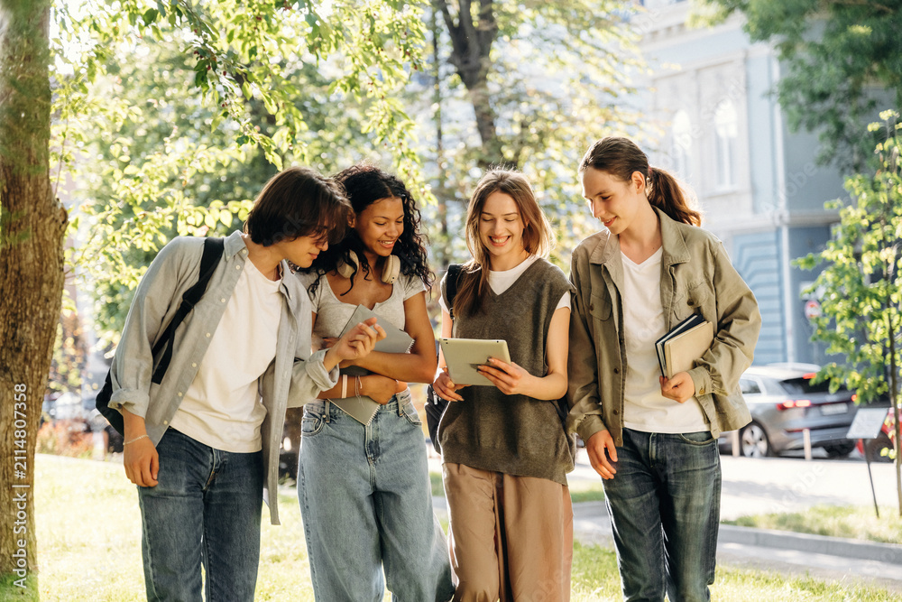 Fototapeta premium Group of four young adults, diverse friends, interacting with a tablet outdoors in a park, surrounded by trees and urban buildings in the background
