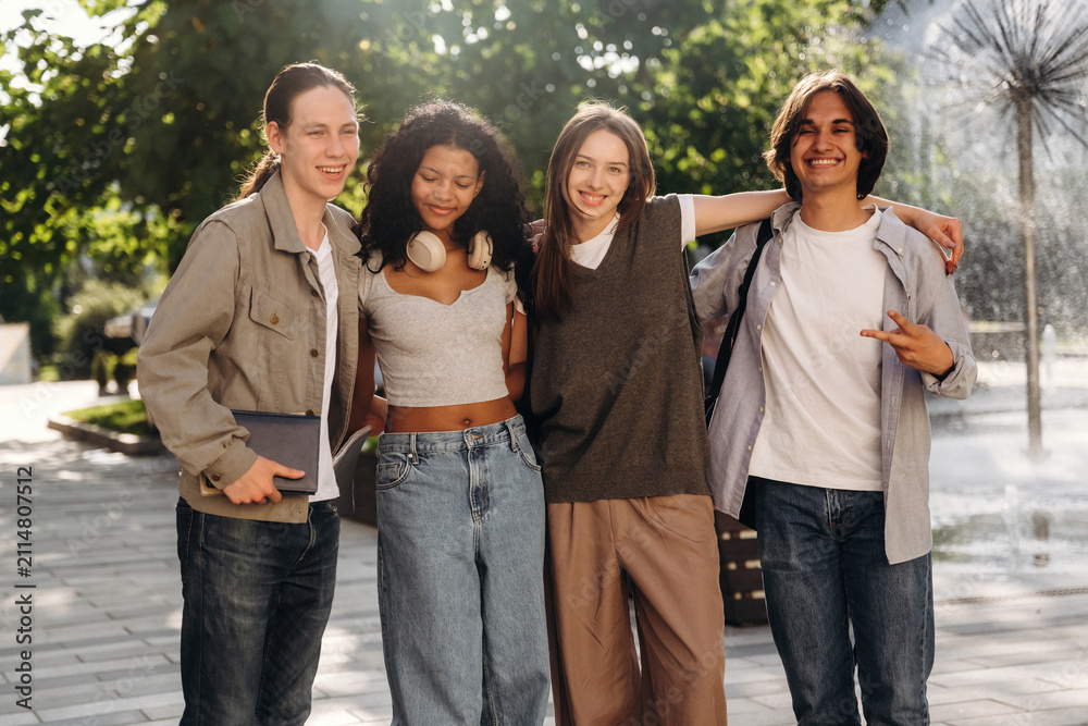 Fototapeta premium Group of four young adults smiling and posing together outdoors near a fountain in a park, wearing casual clothing with greenery in the background