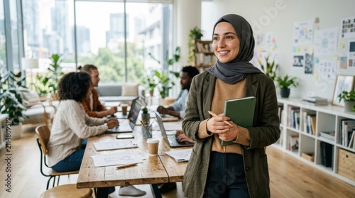 A cheerful young woman wearing a hijab stands in a sunlit creative workspace holding a notebook while her diverse professional team collaborates on new business projects at a large wooden table
