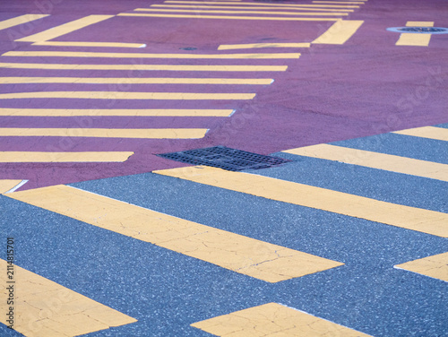 Close-up of a paved urban road intersection with yellow pedestrian crosswalk stripes, grey asphalt, and a reddish-purple surface texture.