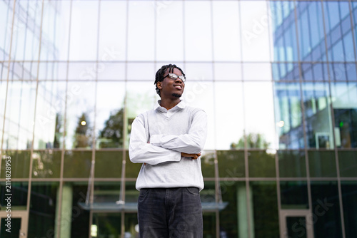 A young African American man with dreadlocks and glasses stands with his arms crossed in front of a modern glass building