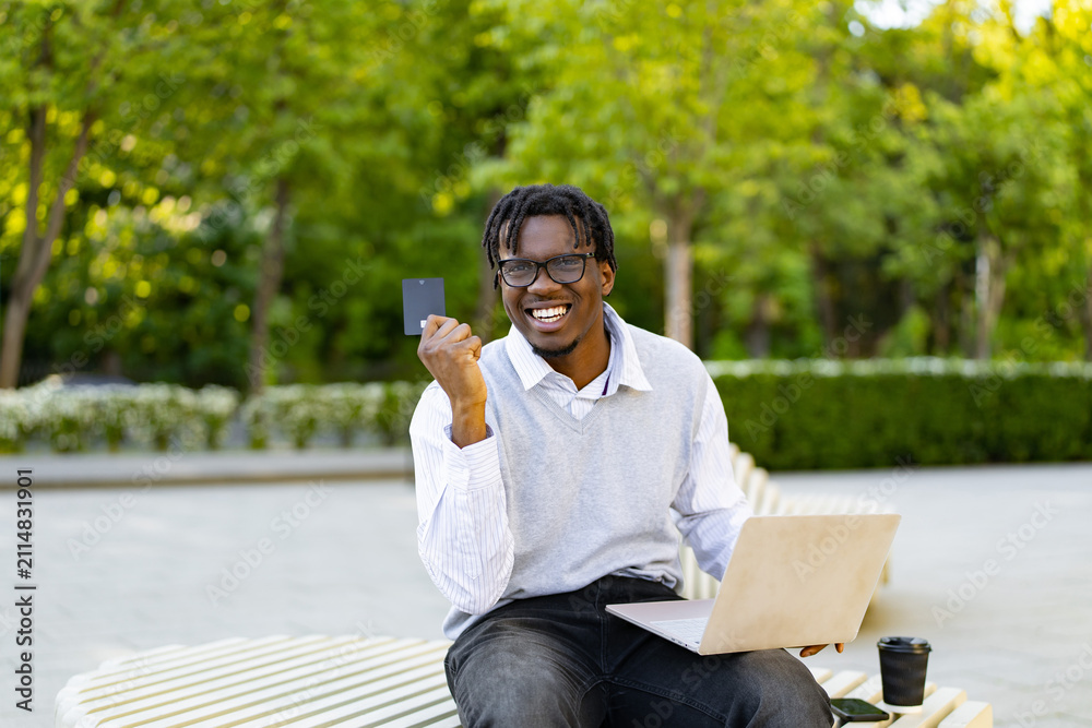 Fototapeta premium A smiling african american man sits outdoors with a laptop and credit card, ready for online transactions