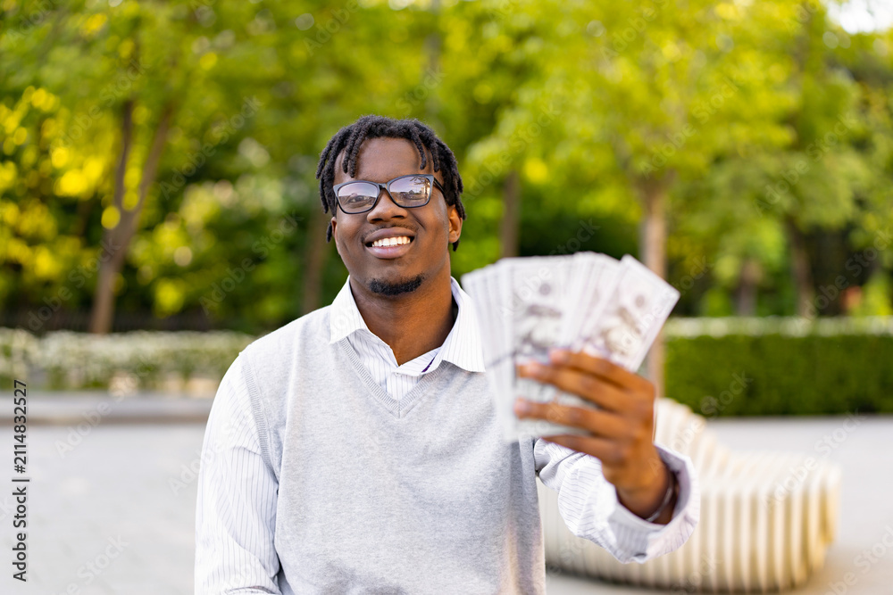 Fototapeta premium A smiling young African American man in glasses holds a fan of US dollar bills in his hand while standing outside in a park