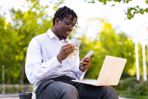 A smiling African American man sits outdoors with a laptop and counts a stack of US dollar bills, possibly managing finances or celebrating a success