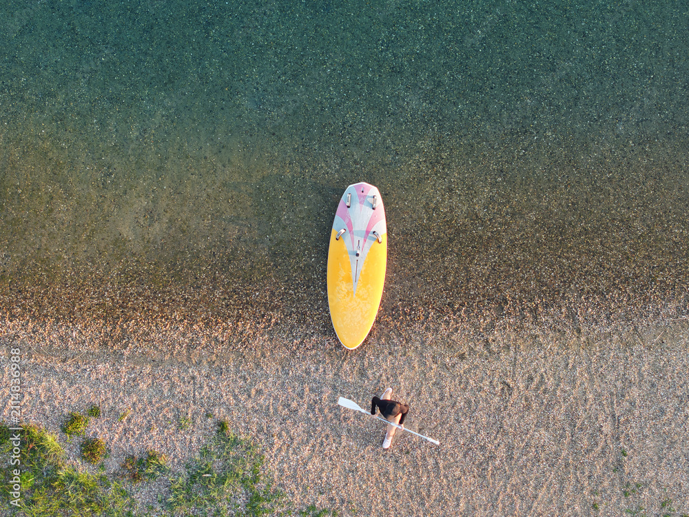 Fototapeta premium Top-down drone view woman carrying paddle beside colorful board on pebbled lakeshore. Clear shallow water, warm sunlight, and graphic composition create calm sunny summer recreation scene