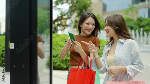 Two happy Asian women with shopping bags use a smartphone to scan a QR code on an interactive digital kiosk screen at an outdoor mall, enjoying retail therapy and modern city lifestyle technology.