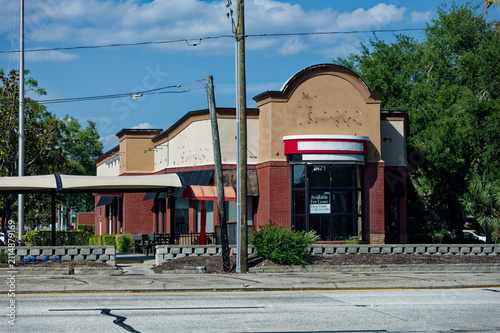 Vacant fast-food restaurant and drive-thru visible from the street with a 'for lease' sign in the window. Empty patio area, and suburban storefront architecture on a sunny day.
