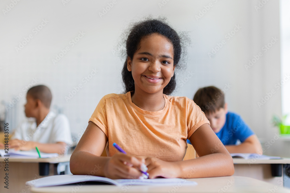 Obraz premium Smiling black schoolgirl writing in open notebook at her desk and looking toward the camera, while diverse classmates study independently in a bright modern classroom behind her