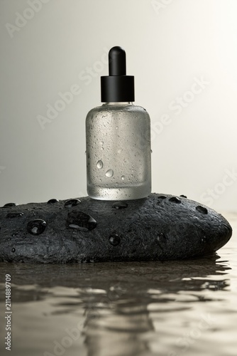Serum bottle on wet stone. Cosmetic skincare product with water droplets on dark rock.