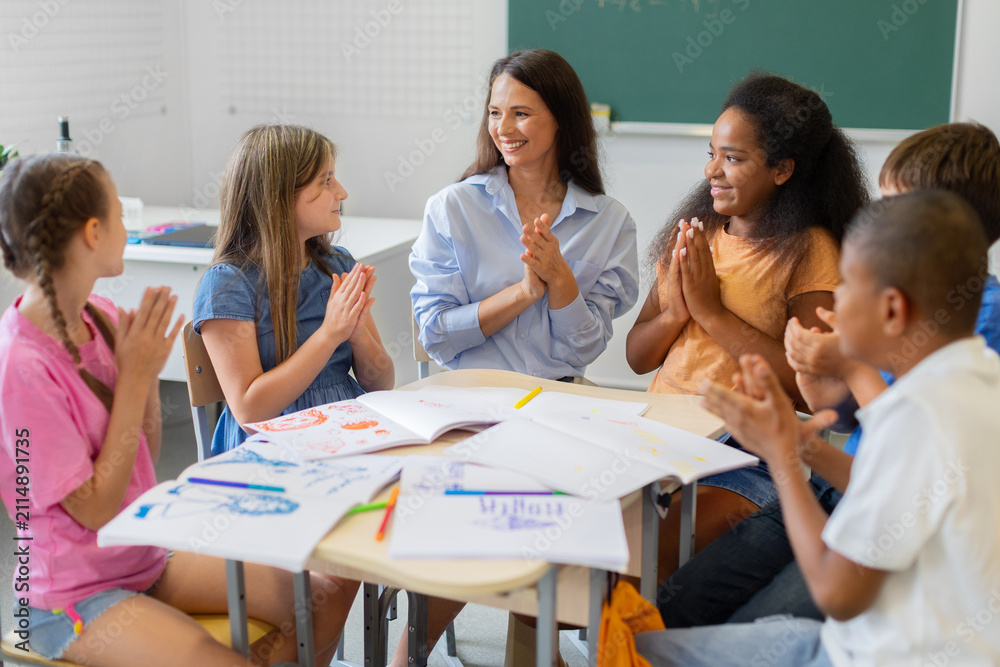 Obraz premium Smiling female teacher and diverse schoolchildren applaud after completing a creative classroom project, celebrating teamwork, achievement, participation, and shared academic success