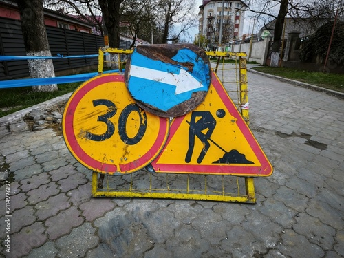 Temporary roadwork warning signs with speed limit and blue directional arrow on cracked city sidewalk beside utility construction trench and fencing