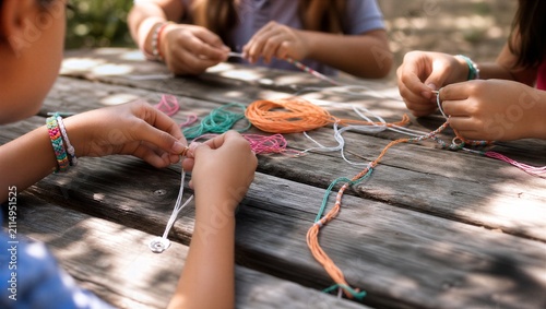 Children sitting at wooden picnic table making colorful friendship bracelets. Summer camp craft activity. Kids braiding thread together outdoors. Friendship concept