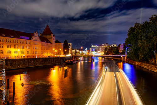 Фотография Night long-exposure view of a Berlin waterway, with a boat's light trail streaking across the water, historic buildings glowing warmly on the left bank, and colorful reflections under a cloudy sky