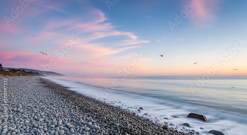 Beach coastline features smooth gray pebbles meeting calm ocean waves under a vibrant pink and blue sunrise.