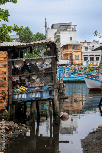 A makeshift wooden stilt shack at the water's edge in a Vietnamese harbour, its shelves holding metal cooking pots. Modern buildings and fishing boats are reflected in the water behind it.