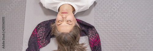 Middle Aged Woman Lying Relaxed With Eyes Closed On Yoga Mat Indoors From Overhead View On Textured Grey Floor Wearing White Shirt And Patterned Scarf Supporting Head During Mindfulness Session.