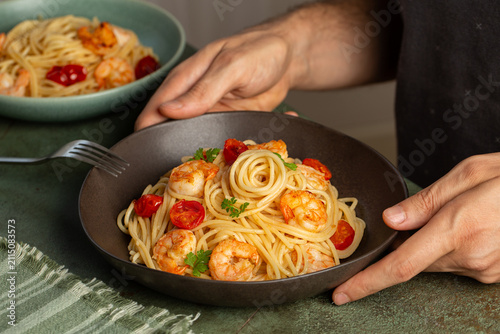 Man hands holding a plate with pasta spaghetti with pan-fried shrimp, cherry tomatoes, butter, fresh parsley. Modern styling on a dark green table.