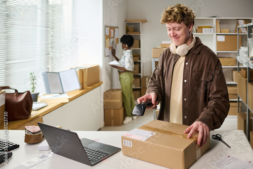 Young adult Caucasian man scanning shipping label on parcel in fulfillment workspace while coworker checking inventory, illustrating ecommerce order processing for retail logistics marketing