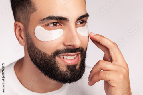 Guy wearing white hydrogel patches under eyes for beauty therapy and dark circles reduction procedure isolated over gray background