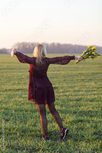 Young blonde woman in long sleeve tiered burgundy ruffled dress standing backwards on green field with open arms and looking back towards meadow and blurred background of trees