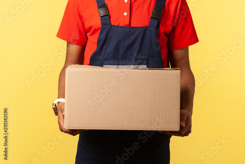 Smiling positive courier wearing blue overalls holding cardboard parcel, delivering orders to address, looking at camera with friendly expression. Indoor studio shot isolated on yellow background.