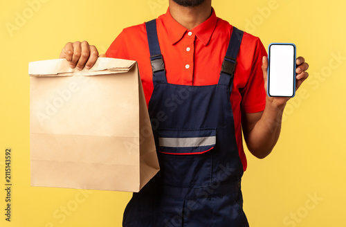 Delivery service mobile app. Deliveryman in overalls showing paper parcel and cellphone with blank display, mock up for express shipping advertise. Indoor studio shot isolated on yellow background.