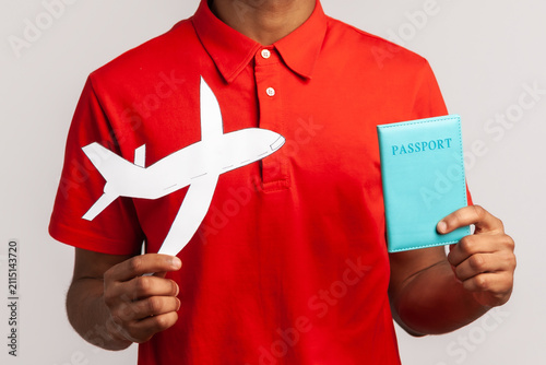 Satisfied man with happy expression wearing red casual style T-shirt, holding passport and paper plane in hands planning vacation abroad. Indoor studio shot isolated on gray background.