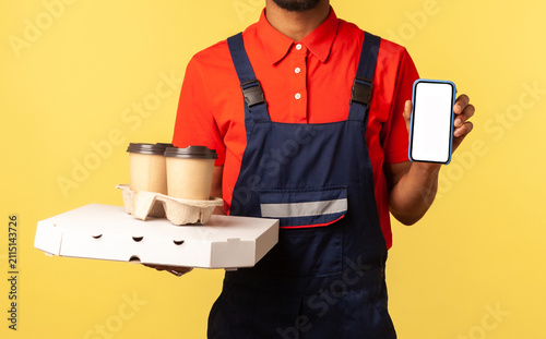 Delivery mobile app, internet advertise. Friendly courier man in overalls holding coffee cups and cellphone with mock up display, online order service. Indoor studio shot isolated on yellow background