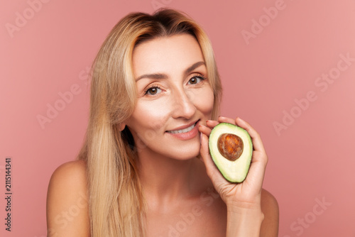 Smiling lady posing with half of ripe avocado near cheek, highlighting moisturizing benefits of natural oils for skin health isolated on pink background