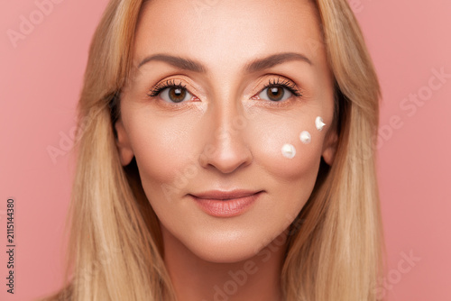 Close-up front view of blonde mature woman with three moisturizer dots applied under her eye looking at camera isolated on pink background