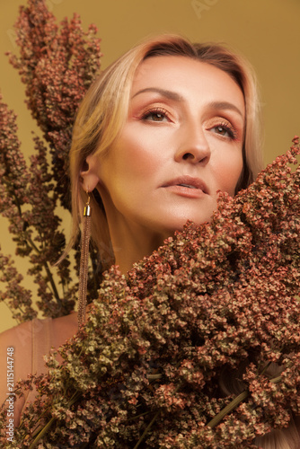 Graceful blonde woman looking up surrounded by dried sorghum plants isolated over yellow studio background