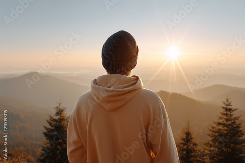 Rear view of young man in hoodie and beanie looking at misty mountain sunset or sunrise