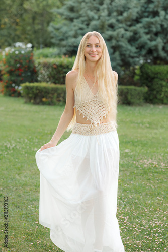 Fotografía A young woman with long blonde hair stands smiling in a white flowing skirt in a