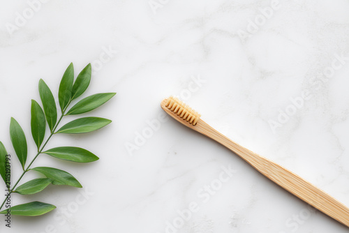Top view of eco friendly wooden bamboo toothbrush next to green plant branch on white marble background