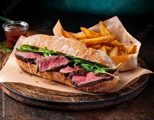 tender medium rare ribeye steak sandwich on baguette alongside fries