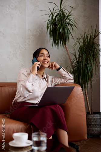 Smiling businesswoman talking on mobile phone while using laptop at hotel reception area.