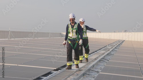 Engineer in safety harness and helmet using radio and tablet while inspecting a large rooftop solar panel installation at a solar power plant.