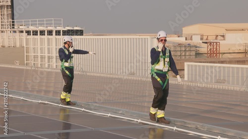 Engineer in safety harness and helmet using radio and tablet while inspecting a large rooftop solar panel installation at a solar power plant.