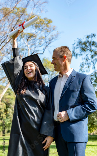 Vertical portrait of a female student in cap and gown raising her diploma alongside a proud middle aged man