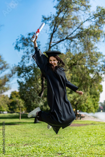 Jumping female graduate celebrating success while holding her diploma scroll high outdoors