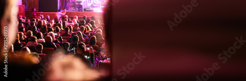 A view of a seated audience in a dimly lit auditorium or theater hall watching a performance on stage under stage lights, illustrating entertainment, events, and culture. Banner with copy space