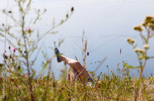 A girl relaxing on a grassy hill overlooking a calm blue lake with a distant boat, framed by wild grass and flowers, illustrating summer vacation, relaxation, and outdoor leisure