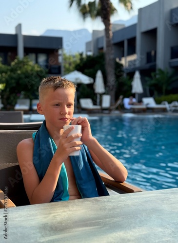 Portrait of a young boy relaxing by a swimming pool. A white plastic cup. Wrapped in a towel after swimming, he enjoys a sunny summer vacation in a tropical resort. Family travel and tourism