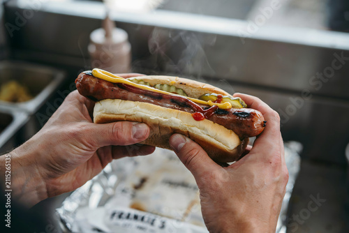 A person holding a steaming hot dog with mustard and ketchup in a bun, prepared inside a food truck kitchen with metallic surfaces visible