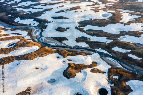 Aerial view of a winding river flowing through a snowy icelandic landscape with patches of brown volcanic earth and melting ice formations
