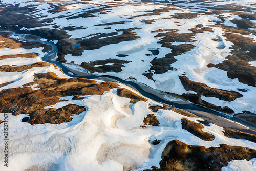 Altitude drone shot of a glacial river winding through a vast snowy valley with patches of exposed dark brown volcanic soil and rocks